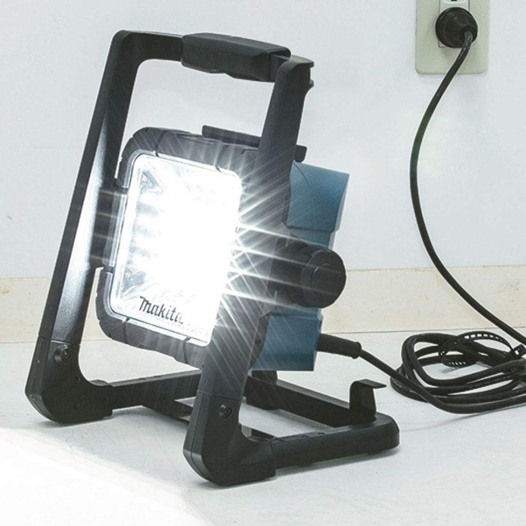 MAKITA LED-valaisin LXT. Kuvassa LED-valaisin LXT oikealta etuyläviistosta valaisin päällä ja yhdistettynä seinäpistokkeeseen virtajohdon avulla.