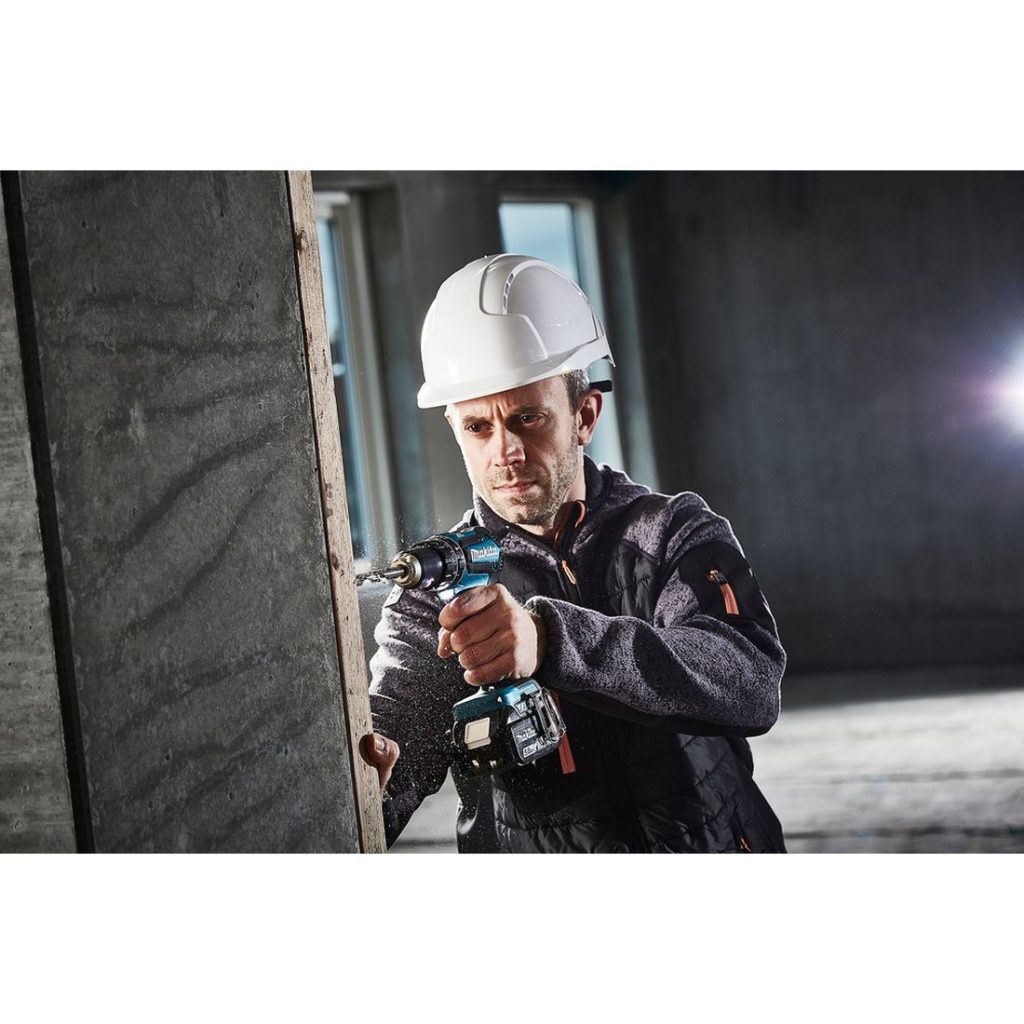 MAKITA Iskuporakone LXT. Kuvassa henkilö poraamassa reikää laudan läpi betoniseinään iskuporakoneen avulla.