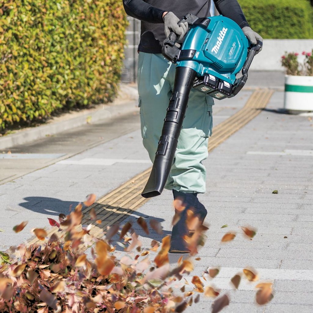 MAKITA Puhallin LXT. Kuvassa henkilö puhaltamassa puun lehtiä kävelykadulla Puhallin LXT:n avulla.