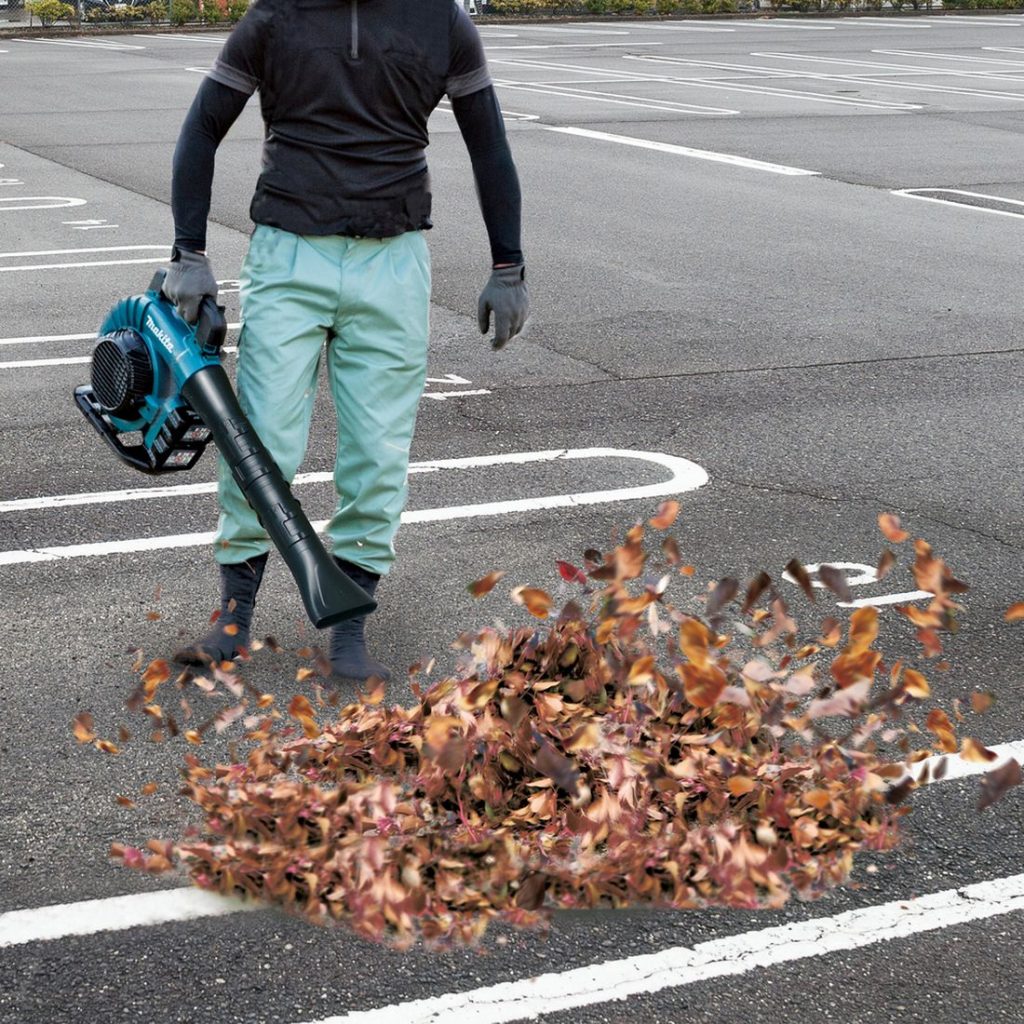MAKITA Puhallin LXT. Kuvassa henkilö puhaltamassa puun lehtiä parkkipaikalla Puhallin LXT:n avulla.