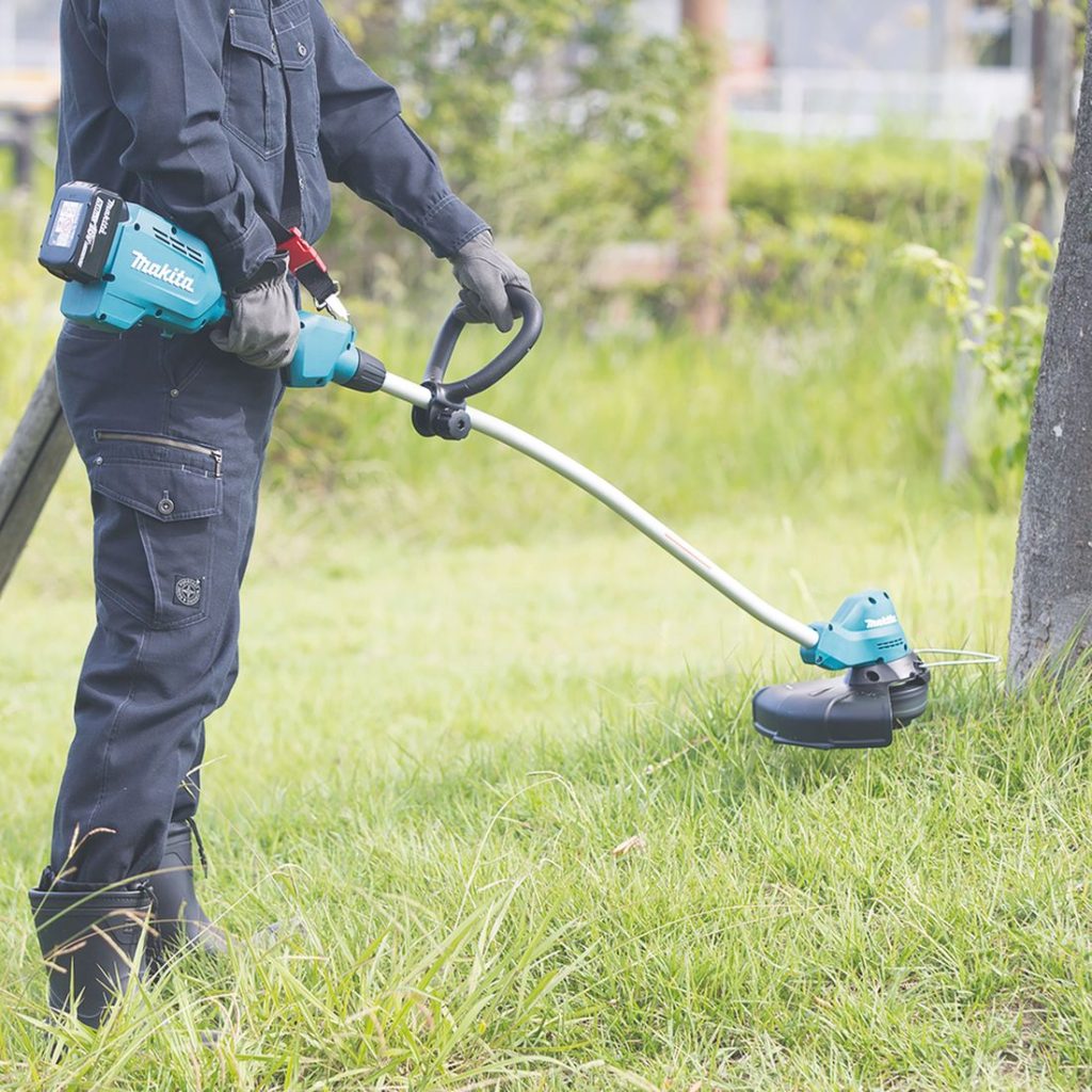 MAKITA Ruohotrimmeri LXT. Kuvassa henkilö leikaamassa ruohoa puun juurien ympäriltä Ruohotrimmeri LXT:n avulla.