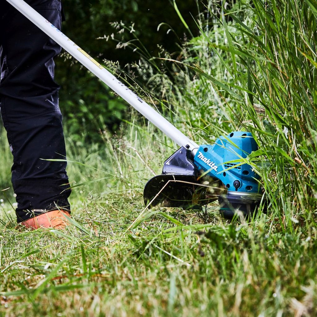 MAKITA Ruohotrimmeri LXT. Kuvassa henkilö trimmaamasssa nurmikkoa pensaan vierestä Ruohotrimmeri LXT:n avulla.