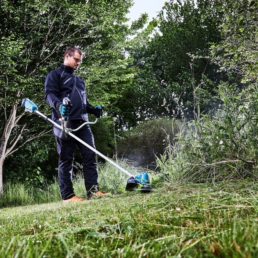 MAKITA Ruohotrimmeri LXT. Kuvassa henkilö trimmaamasssa nurmikkoa pensaan vierestä Ruohotrimmeri LXT:n avulla.