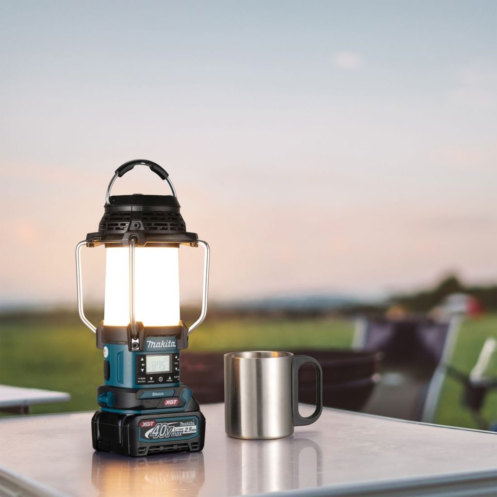 MAKITA Lyhty / Radio / Kaiutin XGT. Kuvassa Lyhty / Radio / Kaiutin XGT ja teräsmuki retkipöydän päällä.