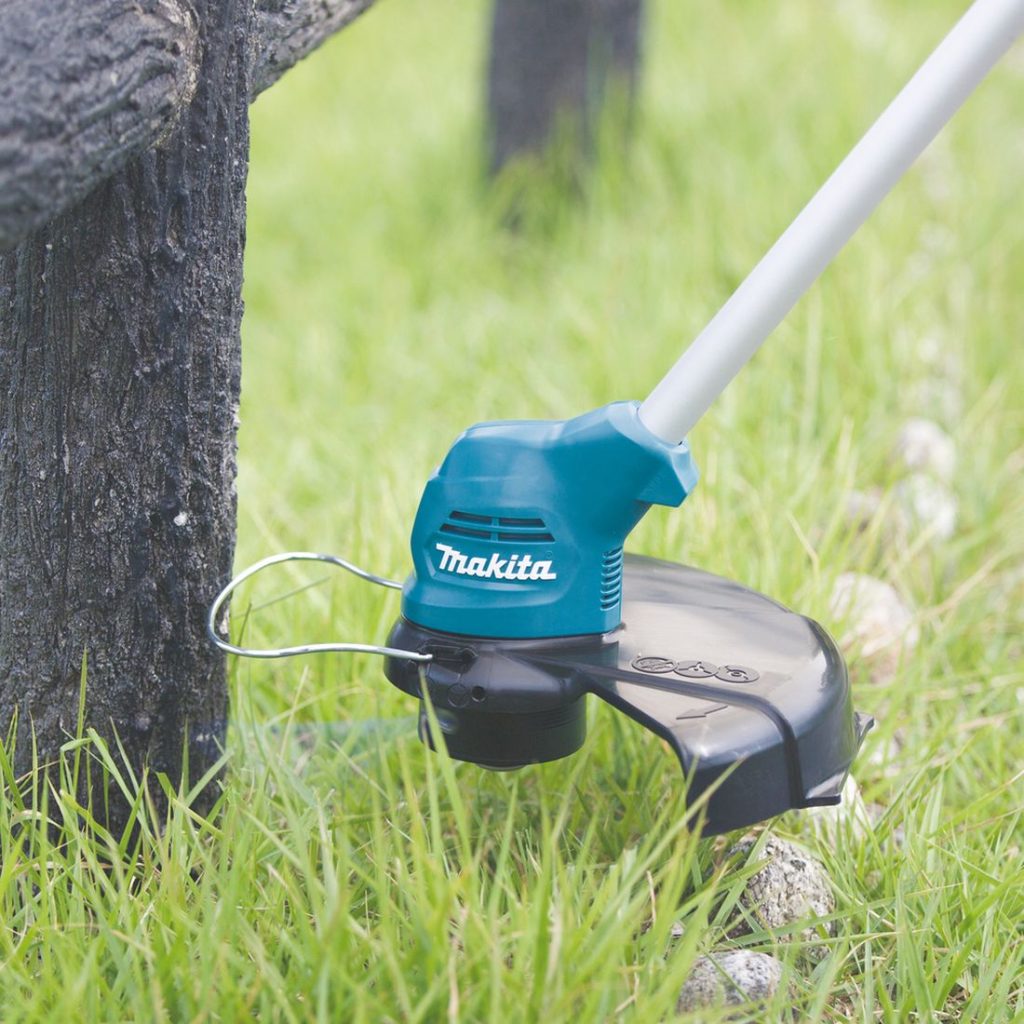MAKITA Ruohotrimmeri CXT. Lähikuvassa henkilö trimmaamassa nurmikkoa aidan tolpan vierestä Ruohotrimmeri CXT:n avulla.