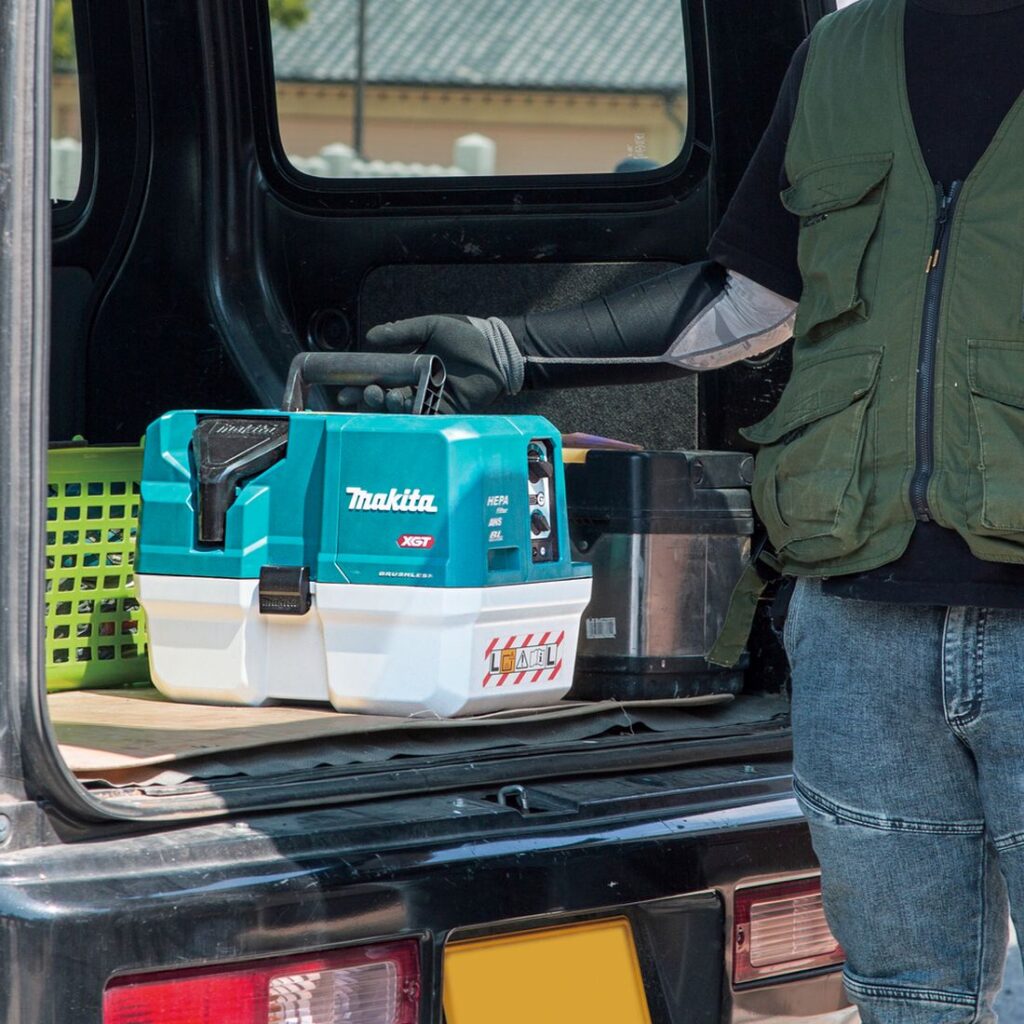 MAKITA Akkuimuri XGT. Kuvassa henkilö nostamassa akkuimuria auton takakontista taittuvaa kahvaa hyväksi käyttäen.